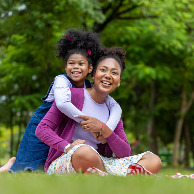Mother Is Playing Piggyback Riding And Hugging With Her Young Daughter While Having A Summer Picnic In The Public Park